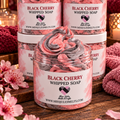 Four jars of Miss Jules Melts Black Cherry Whipped Soap arranged on a wooden surface, with one jar open in front to show the swirled whipped soap inside. The setting is styled with pink cherry blossoms and a cozy knit blanket.