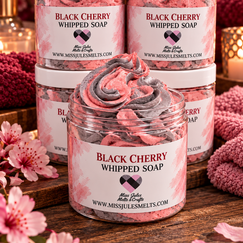 Four jars of Miss Jules Melts Black Cherry Whipped Soap arranged on a wooden surface, with one jar open in front to show the swirled whipped soap inside. The setting is styled with pink cherry blossoms and a cozy knit blanket.