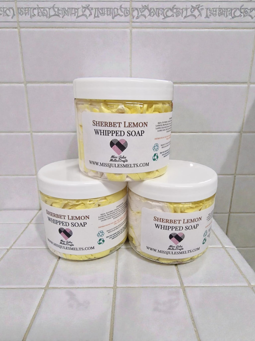 Three jars of Sherbet Lemon Whipped Soap stacked in a pyramid arrangement on a white tiled surface against a matching tiled background.