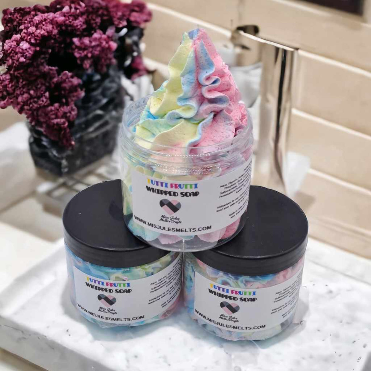 Three jars of Luvy Brubro Whipped Soap stacked on a marble tray in a bathroom setting, with a colorful whipped soap product on top and two black-lidded jars below.